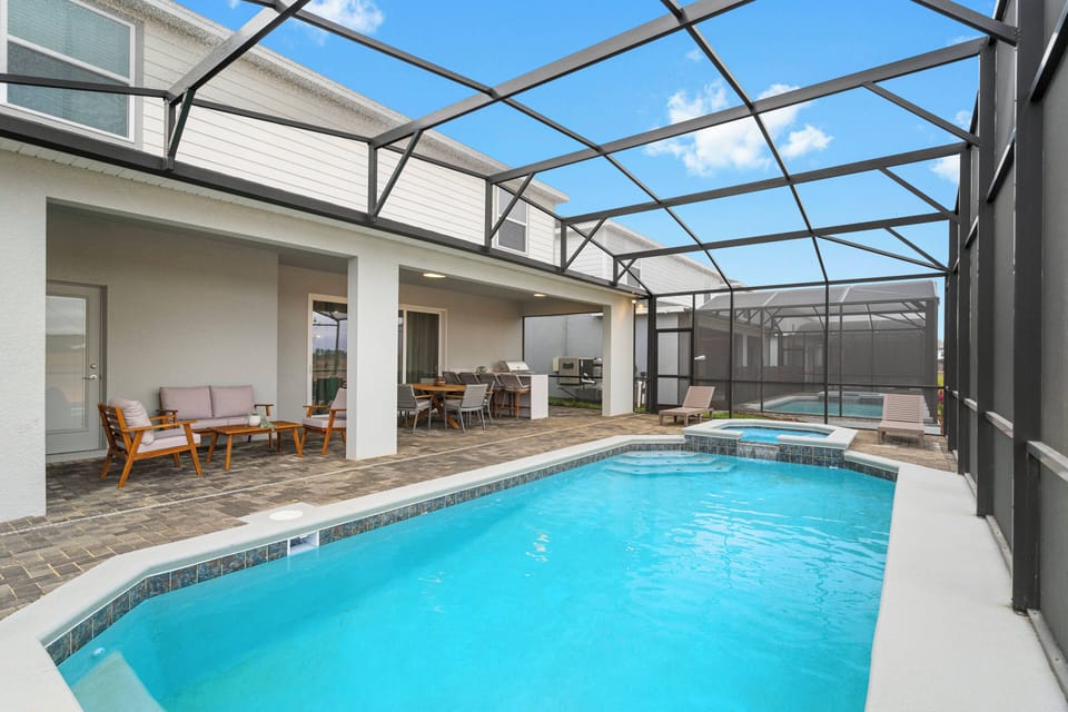 Enjoy a bright private screened pool with a covered patio, outdoor seating, dining table, grill, and lounge chairs. The open sky, modern deck, and relaxing water views create a perfect space for sunny Florida vacation days.