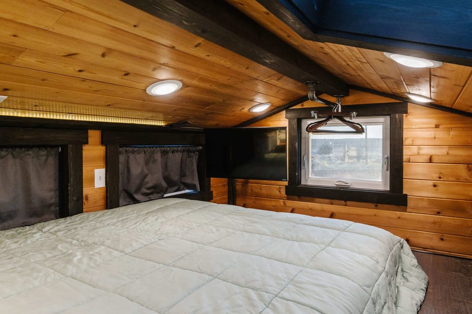 Cozy queen loft bedroom with warm wood ceilings, skylight, and soft lighting. Comfortable mattress and thoughtful layout create a quiet, relaxing sleep space in this charming tiny home retreat.