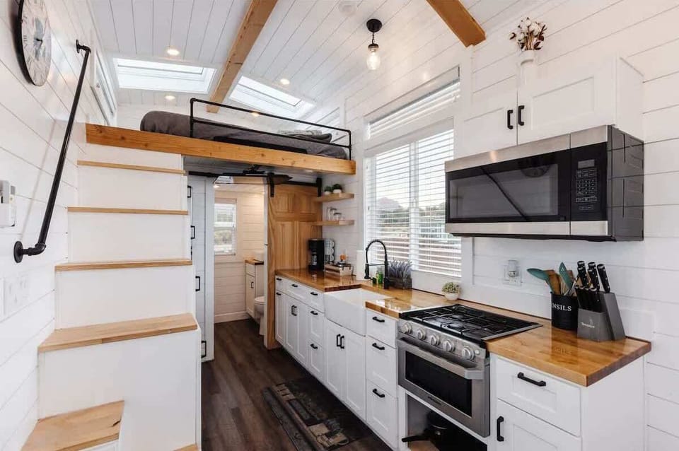 Bright full kitchen with gas range, microwave, farmhouse sink, and butcher block countertops. Stairs lead to the loft bedroom, maximizing space in this modern tiny home near Zion.