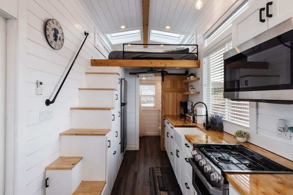 Spacious tiny home kitchen featuring a full gas range, stainless appliances, open shelving, and smart storage beneath the staircase for a functional, comfortable stay.