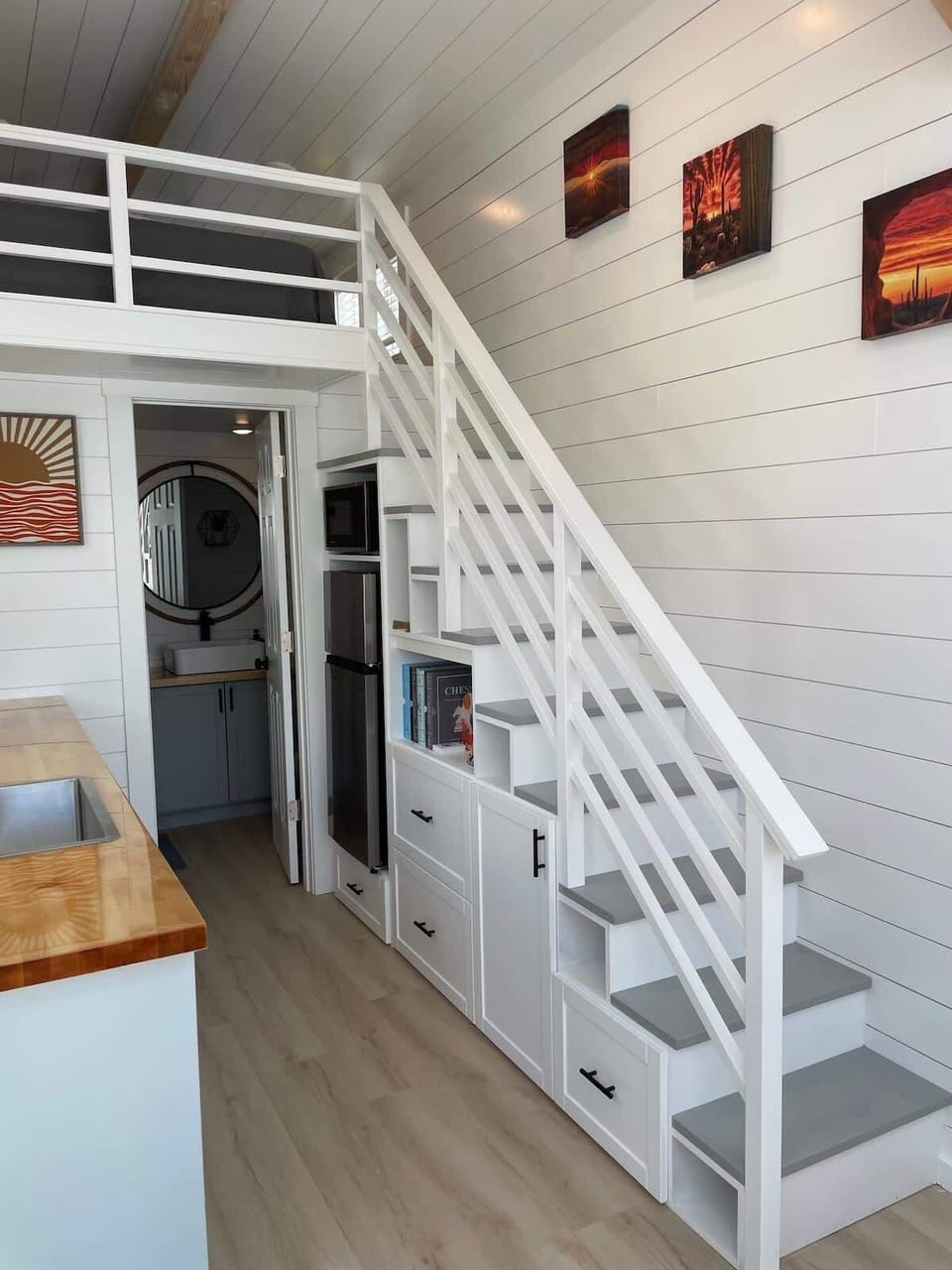 Open living area with vaulted ceilings, loft staircase, smart TV, and modern finishes throughout. Bright white interior and large windows create an airy, tiny home retreat perfect for relaxing after an adventure.