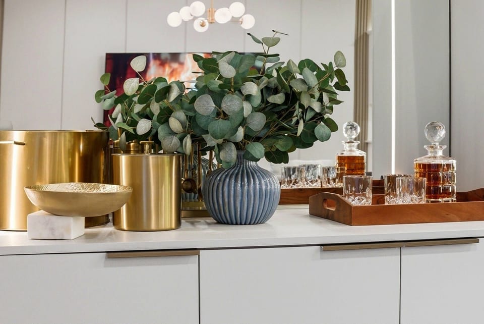 Discover a beautifully arranged console table featuring a textured blue vase with lush eucalyptus, elegant gold accents, and crystal glassware. The warm lighting creates a sophisticated atmosphere, inviting you to relax in style.