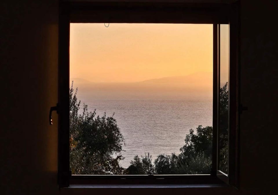 Sunset view toward the sea framed by the window, with olive trees in the foreground.