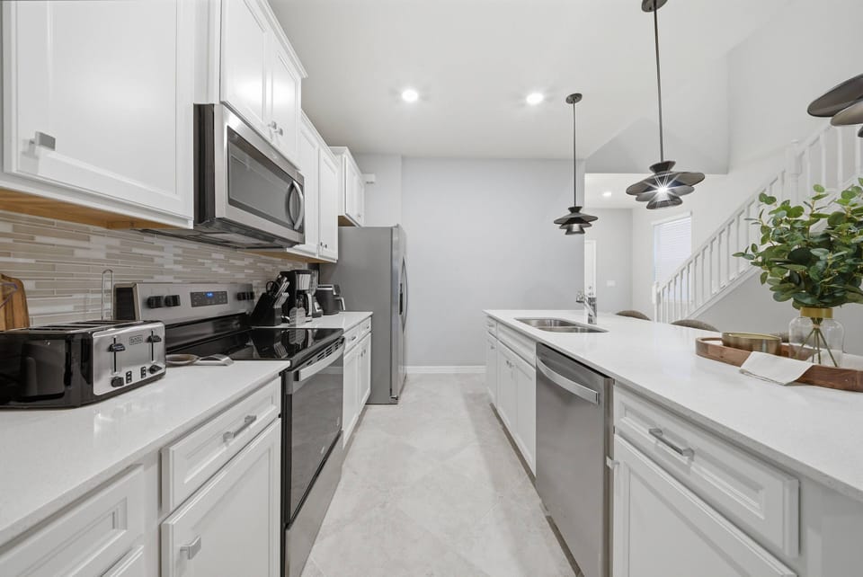 Discover this chic kitchen featuring white cabinetry and sleek stainless steel appliances. The airy design, brightened by pendant lighting, sets the perfect mood for cooking or savoring your morning coffee.