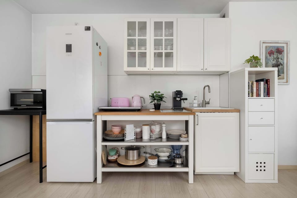 A compact kitchen corner with white cabinets and open shelving. Small appliances and jars are neatly arranged. The space feels functional and well-organized.