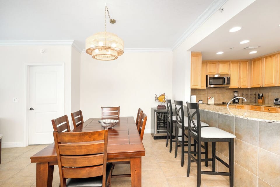 Your elegant dining space features a beautiful chandelier above warm wood table seating six, flowing into a fully equipped kitchen with breakfast bar.