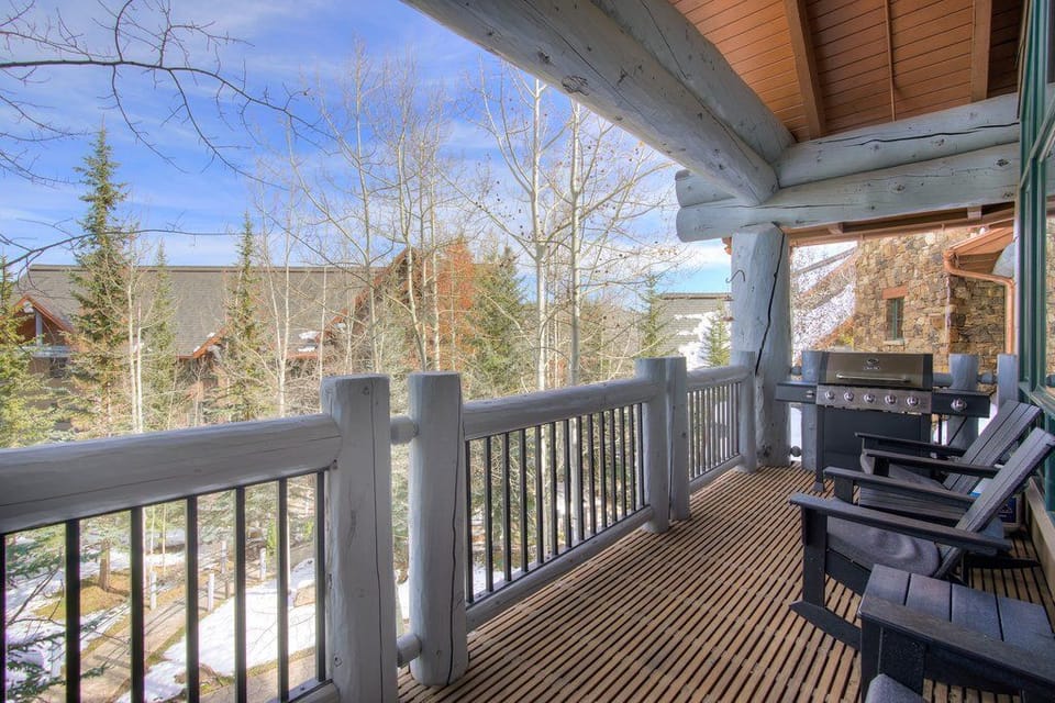 Balcony with wood flooring, outdoor chairs, a barbecue grill, surrounding trees, and nearby buildings.