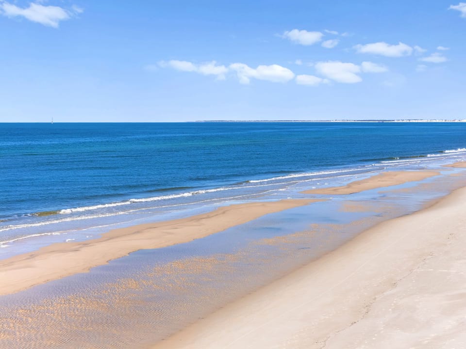 Pristine sandy beach stretches along crystal-clear blue waters under a bright sky with gentle waves lapping the shore.