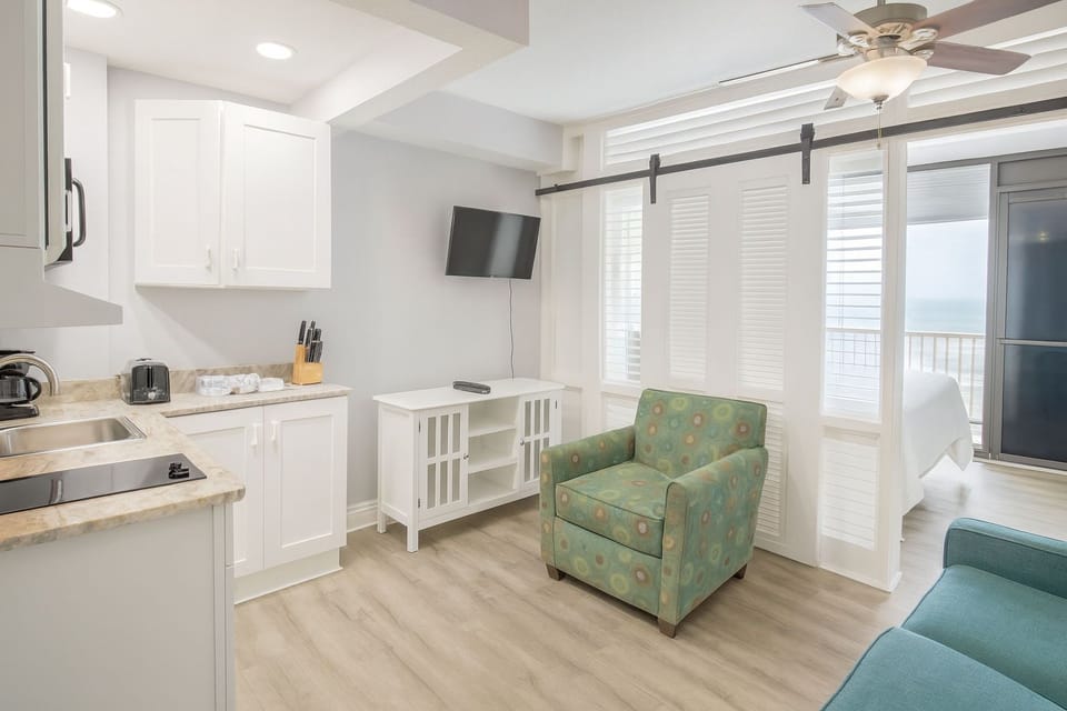 A modern kitchenette area combined with seating area in the Oceanfront Deluxe Studio, featuring sleek white cabinetry and cheerful decor, perfect for gathering during your beach getaway.