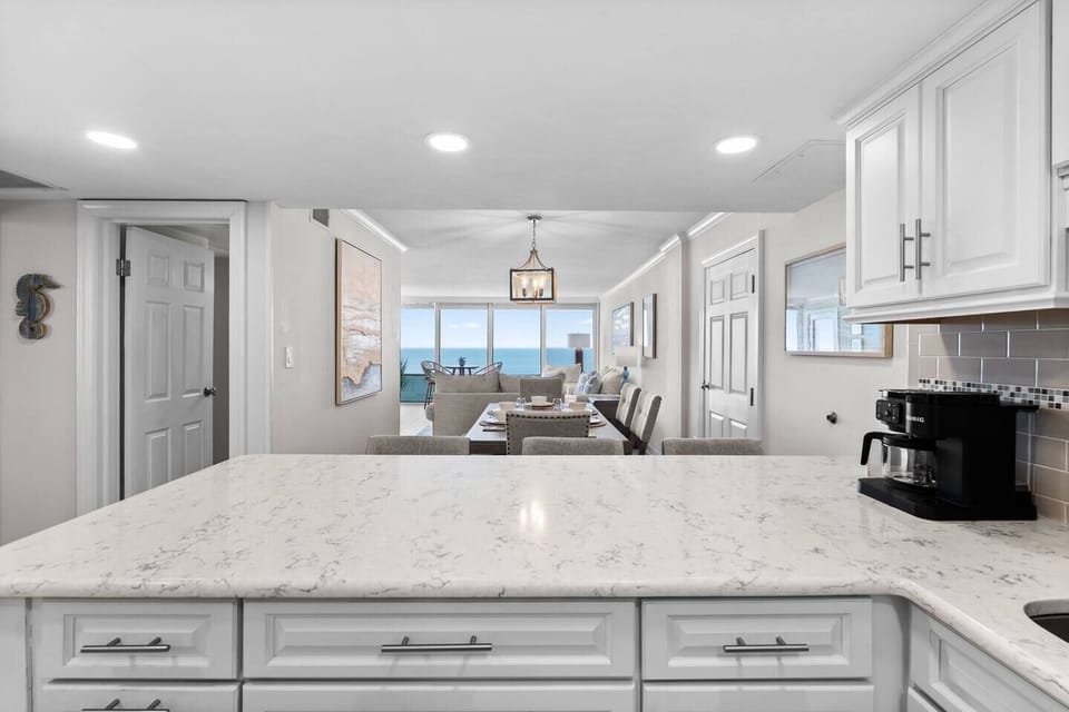 Bright, open kitchen with quartz countertops flowing into the dining area and ocean views beyond.