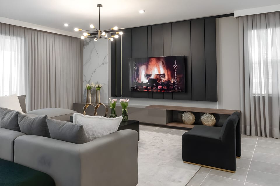 This stylish living room features soft gray tones, sleek furnishings, and a cozy fireplace on a modern media wall. Large windows with light curtains fill the space with natural light, creating a warm atmosphere for your.