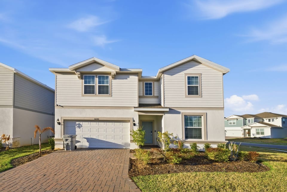 Discover a charming home with light gray siding and a welcoming porch, surrounded by lush landscaping. The inviting brick driveway warmly beckons you to explore the cozy comfort inside.