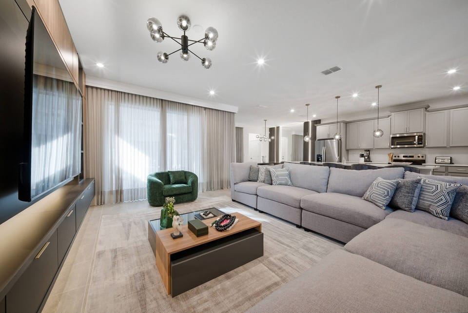 Enjoy the cozy ambiance of this spacious living area, complete with a plush sectional sofa, a stylish coffee table, and large windows dressed in soft curtains that fill the room with warm, natural light for your relaxation.