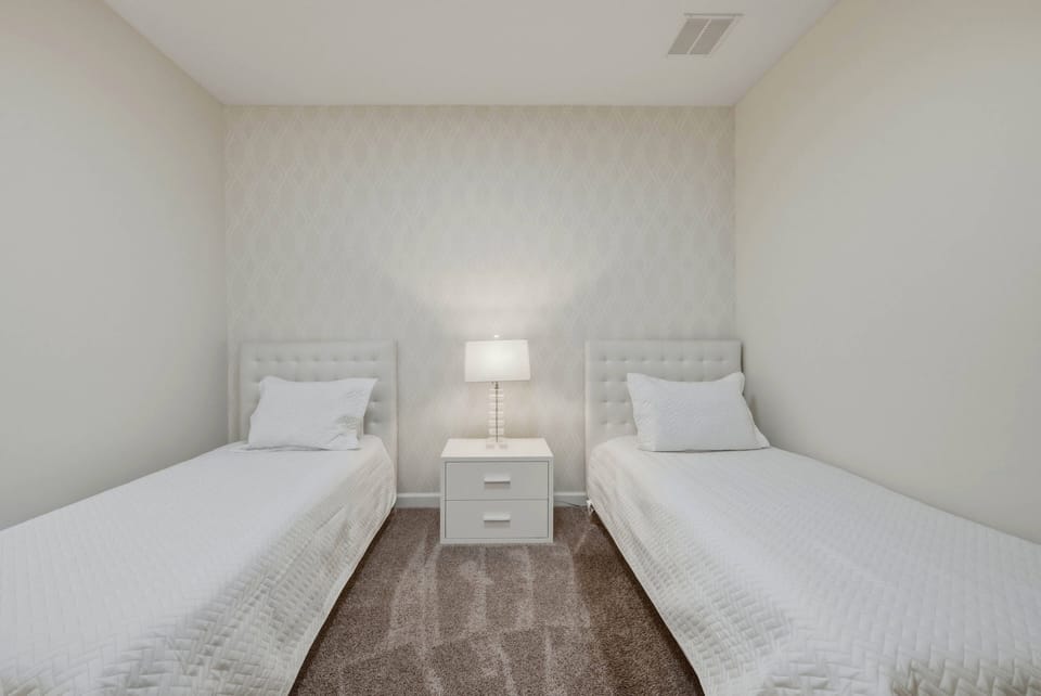 This cozy bedroom offers two neatly arranged twin beds with crisp white linens, complemented by soft neutral wall tones. A stylish bedside table with a modern lamp enhances the warm and inviting atmosphere for restful nights.