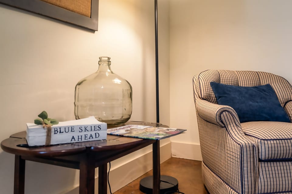 Suite #1 – Cozy Corner: A charming reading nook featuring a classic striped armchair with a plush accent pillow, warm floor lamp lighting, and thoughtful details that invite you to relax and unwind.