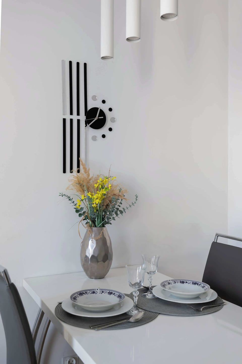 A close-up of a white dining table set with elegant plates, glassware, and a silver vase filled with yellow flowers. An artistic black wall clock with vertical bars and circular hour markers hangs above the table.