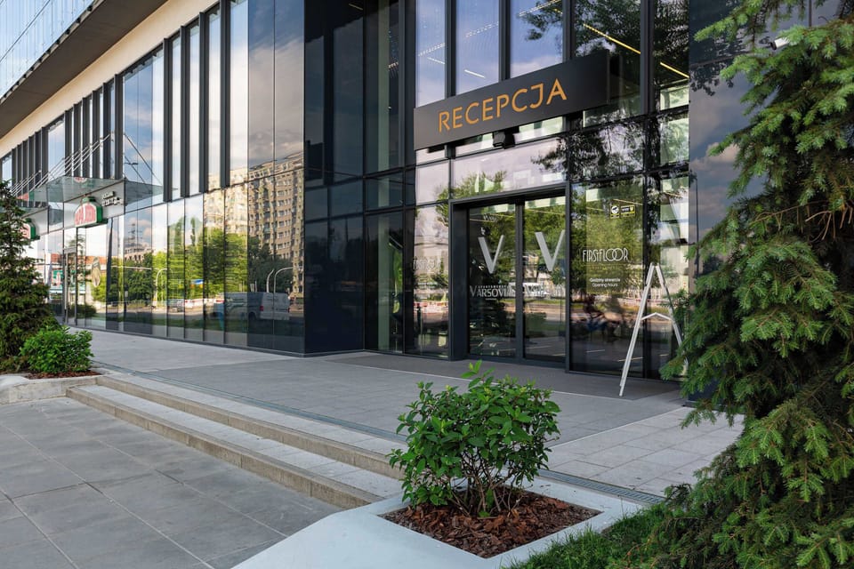 The exterior entrance of the building features a large glass facade with a prominent black sign above the door that reads "RECEPCJA." Neatly manicured greenery and a paved walkway lead guests toward the main revolving or sliding glass doors