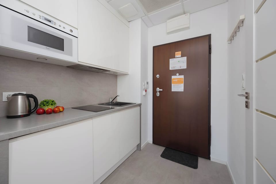 A functional white kitchenette equipped with a built-in microwave, an induction hob, and a dark sink. The brown entrance door is visible at the end of the short hallway next to the kitchen counter.
