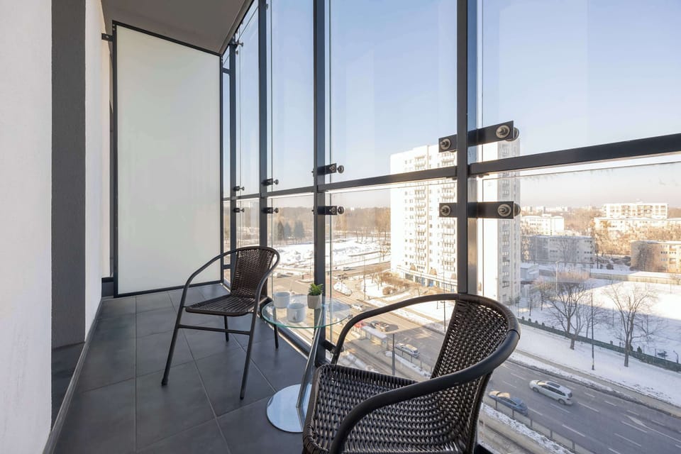 A spacious, glass-enclosed balcony features two dark wicker chairs and a small glass bistro table. The elevated view overlooks a city street and snow-covered grounds during a bright, sunny day.