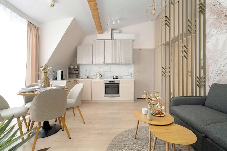 An open-plan living and dining area under slanted ceilings. A sofa, dining table, and kitchen are arranged in one cohesive space. Light colors make the room feel spacious.