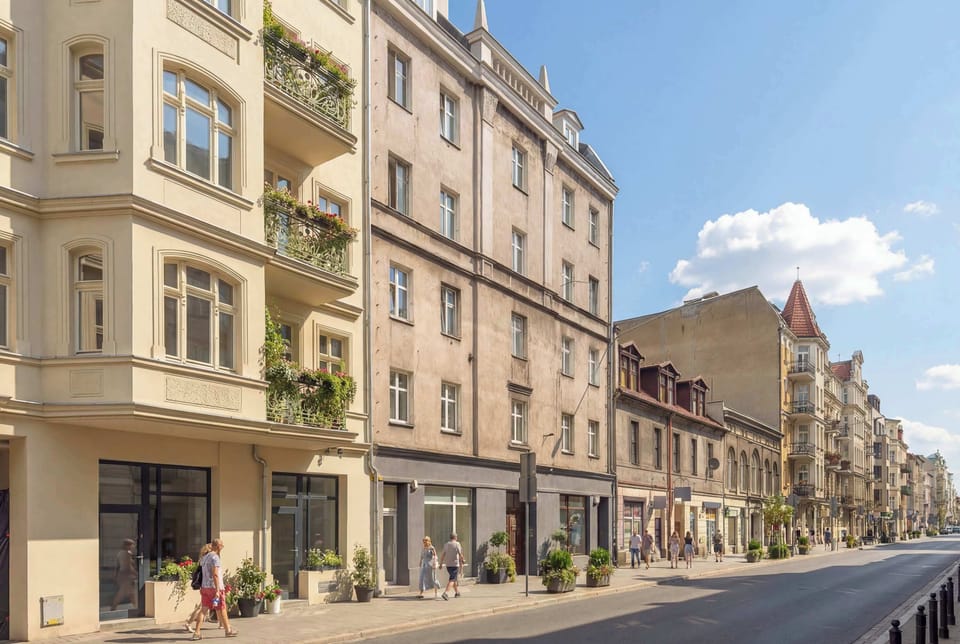 An exterior street view with classic multi-story buildings lined along the road. The architecture features light facades and large windows. The street looks calm and urban.