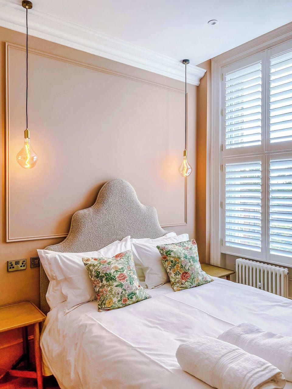 A serene bedroom with padded headboard, floral pillows, and warm pendant lights. Neutral tones and plantation shutters create a calm, welcoming retreat filled with natural light.