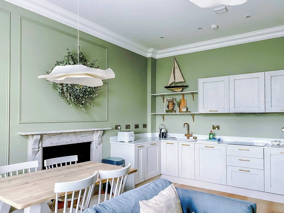A bright kitchen with white cabinetry, gold handles, and sink with faucet. Open shelves with coastal décor, kettle, and sailboat accents flow into a warm dining space with wooden table and white chairs.