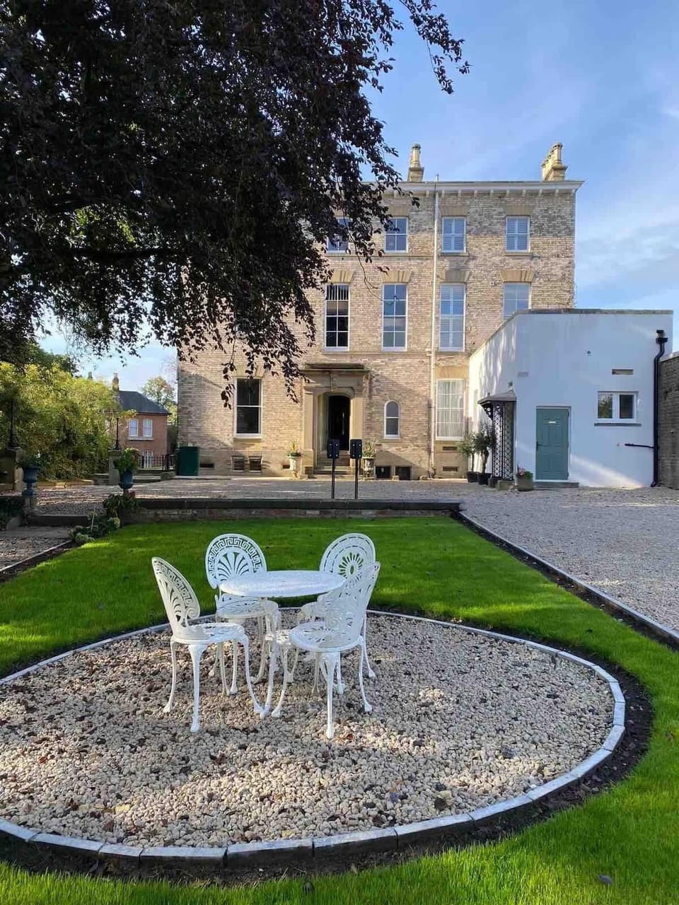 Elegant outdoor courtyard with a circular gravel seating area framed by lush grass. A white metal table and chairs invite relaxation, set against a grand stone building with tall windows and ornate details, with a peaceful garden.