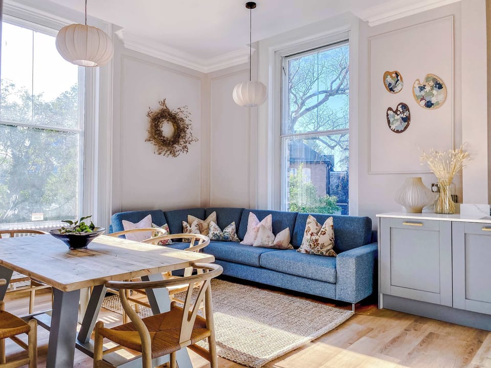 A bright living and dining space with a light blue sectional sofa, decorative pillows, and natural wood table. Pendant lights, wall art, and soft flooring create a warm, welcoming retreat filled with natural light