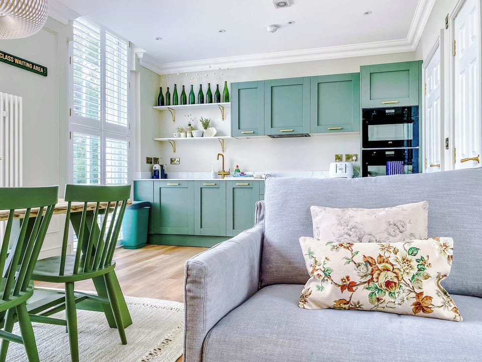 A bright kitchen with sage cabinets, white counters, and built‑in oven and microwave. Open shelves, green accents, and natural light flow into a cozy dining and lounge space with soft seating.