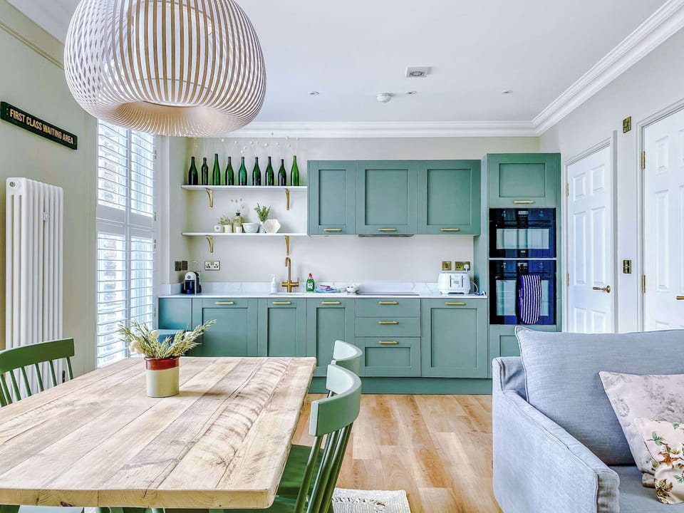 Bright and inviting living room with a cozy gray sofa accented by decorative pillows. Large windows with white shutters fill the space with natural light, while wooden floors and green cabinetry nearby create a stylish open‑plan flow between kitchen,