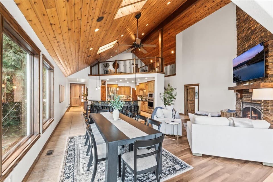 Bright open great room with 10-seat dining table, vaulted wood ceiling, lit fireplace, and forest views