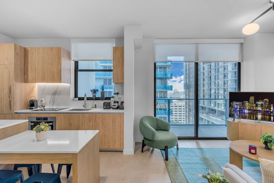 Cook and relax in this stylish open-plan living space. Features a modern kitchen with light wood cabinetry and a spacious island, seamlessly flowing into a bright living area with a balcony overlooking the city skyline