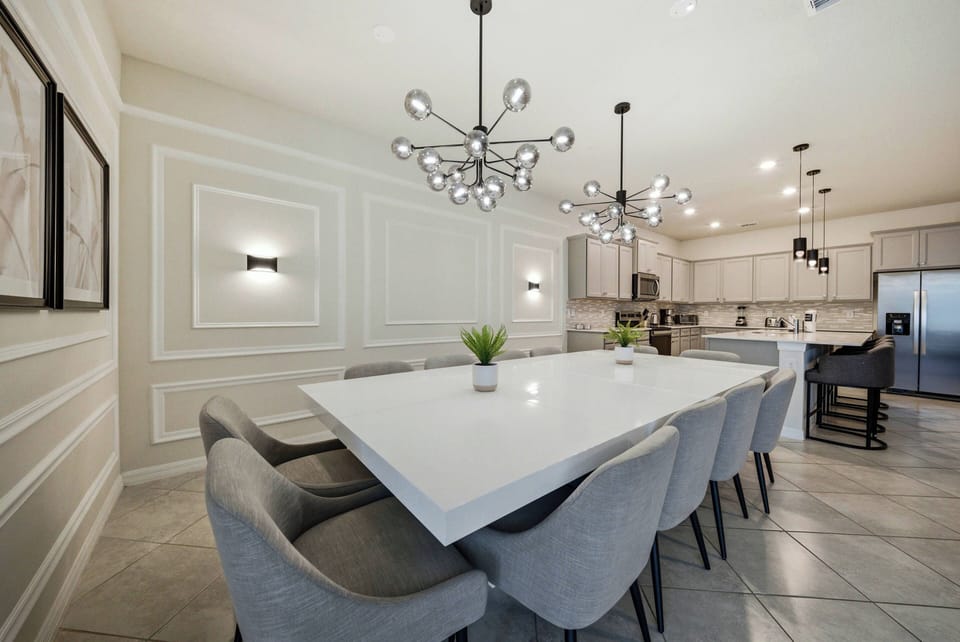 Enjoy meals and gatherings in this stylish dining area, featuring a sleek white table and plush gray chairs. Modern light fixtures and elegant wall detailing set a sophisticated atmosphere, ideal for entertaining your guests.