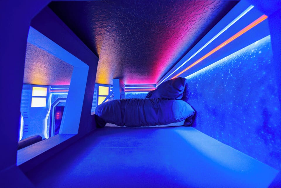 Experience a uniquely designed nook with vibrant blue and neon accents that energize the atmosphere. Relax in the cozy bed against a textured wall, while whimsical lighting adds a playful touch to your stay.