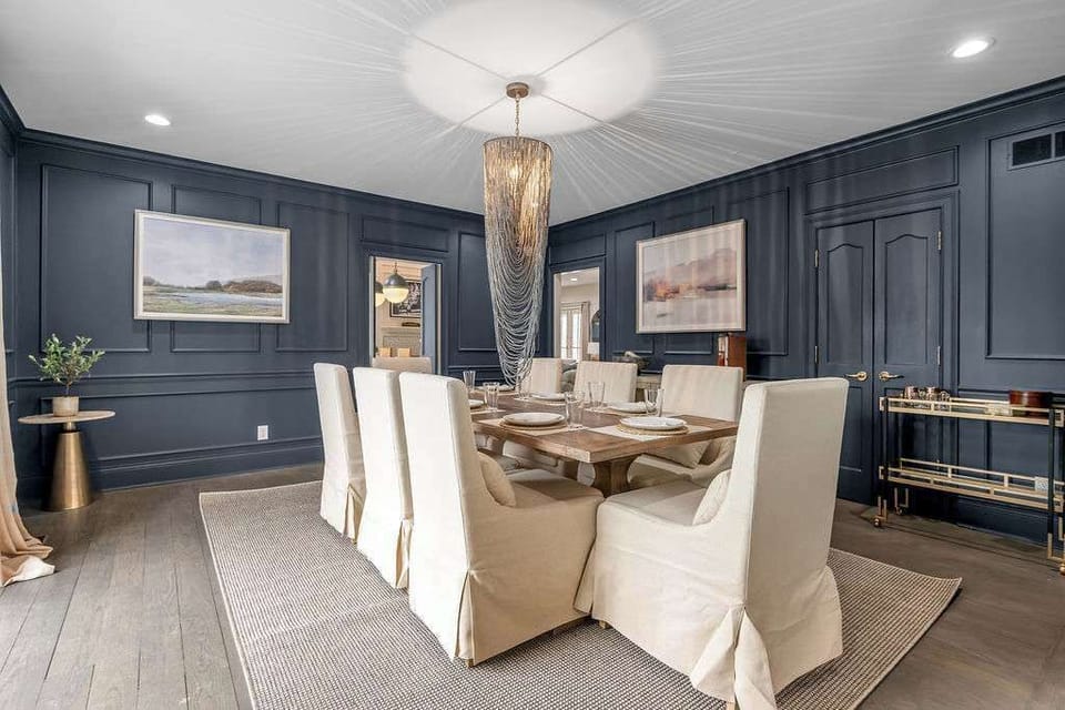 Gather around the long table for a real sit-down dinner — navy walls, crystal chandelier, white slipcovered chairs, room for everyone. The room earns the meal.