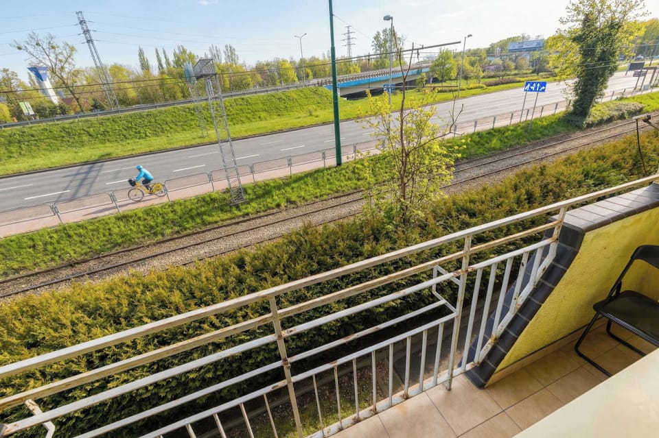 The sunny balcony offers a refreshing view of green trees and a nearby road with a safety railing.
