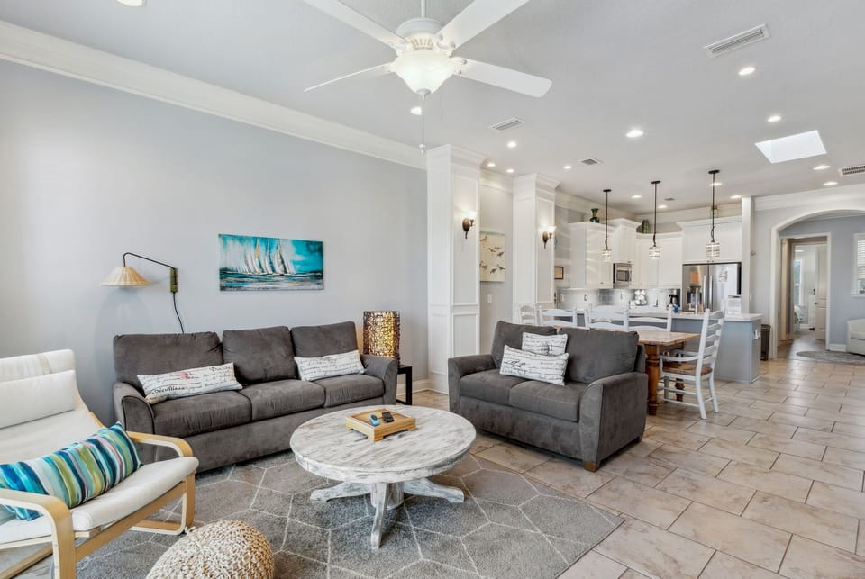 Relax in your spacious living room with plush gray sofas and coastal artwork, flowing seamlessly into a modern kitchen perfect for gathering.