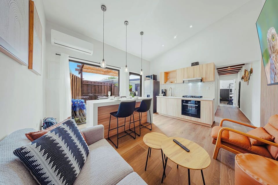 Open-concept living space with sleek kitchen, cozy lounge, and sunlit patio. Light wood finishes, pendant lighting, and warm accents create a modern, inviting atmosphere perfect for relaxing or entertaining.