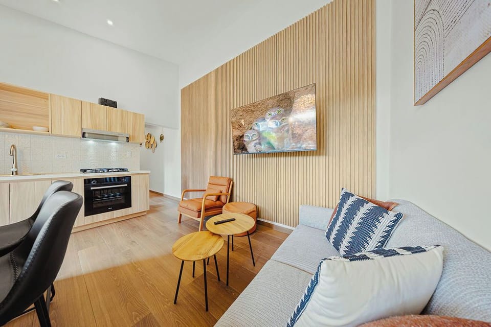 Open-concept living space with sleek kitchen, cozy lounge, and sunlit patio. Light wood finishes, pendant lighting, and warm accents create a modern, inviting atmosphere perfect for relaxing or entertaining.