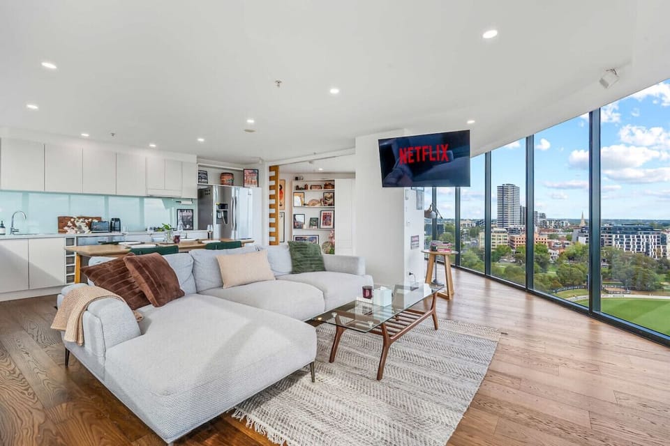 Generous open-plan living space with a large sectional sofa, wall-mounted smart TV with Netflix, and a fully equipped kitchen visible in the background. Floor-to-ceiling windows look out over a cricket oval and the surrounding suburbs.