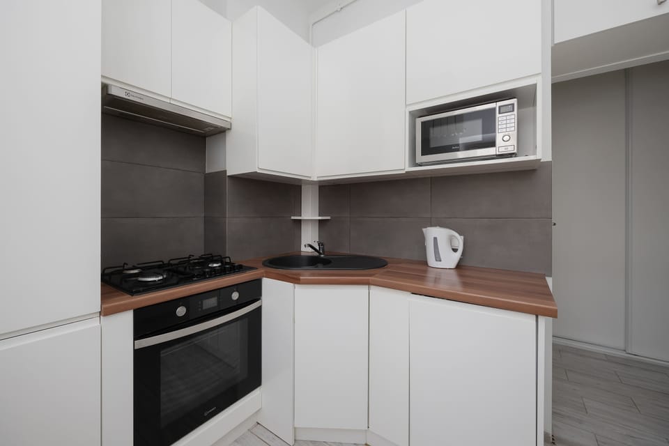 A modern kitchenette displays white cabinetry paired with a dark countertop and a built-in oven. A microwave is tucked into an upper shelf, and a white electric kettle sits ready on the counter.