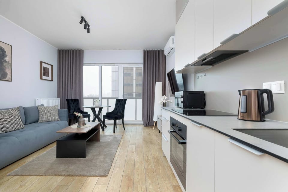A perspective view from the kitchen area toward the living space and balcony. White cabinetry contrasts with warm wooden flooring. The open layout enhances the sense of continuity throughout the studio.