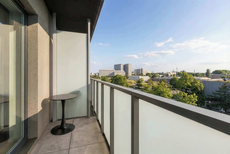 A balcony with glass railings overlooking the city skyline. A small table is placed outside, creating a pleasant spot to enjoy the view. The perspective highlights nearby buildings and open sky.