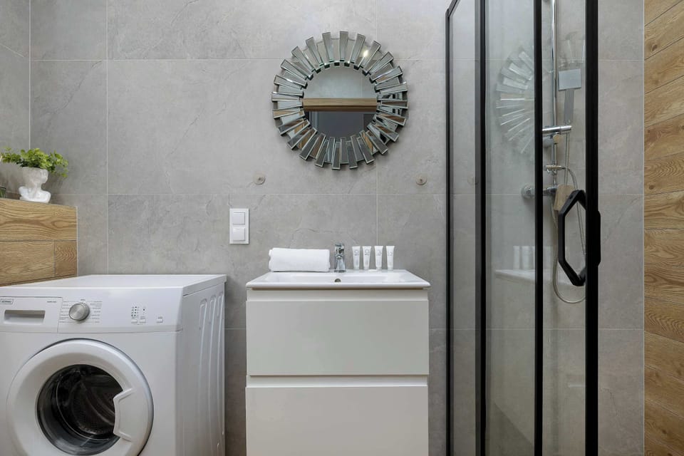 A bathroom corner featuring a washing machine next to a vanity unit and shower cabin. A decorative round mirror hangs above the sink. Practical layout ensures comfort for longer stays.