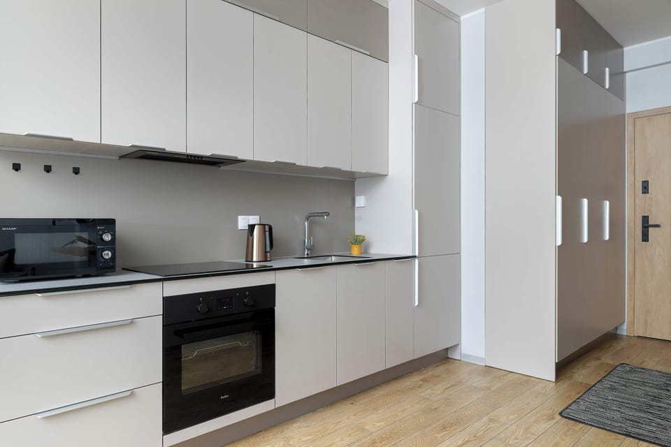A modern kitchenette with white cabinets, built-in oven, and induction cooktop. The countertop includes small appliances and offers plenty of preparation space. Clean lines and neutral colors emphasize a contemporary style.