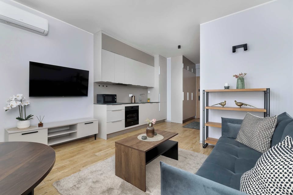 An open-plan living room with a wall-mounted TV and a sleek kitchenette in the background. A wooden coffee table sits in front of the sofa, and modern white cabinetry lines the kitchen wall. The space feels airy and well-organized.