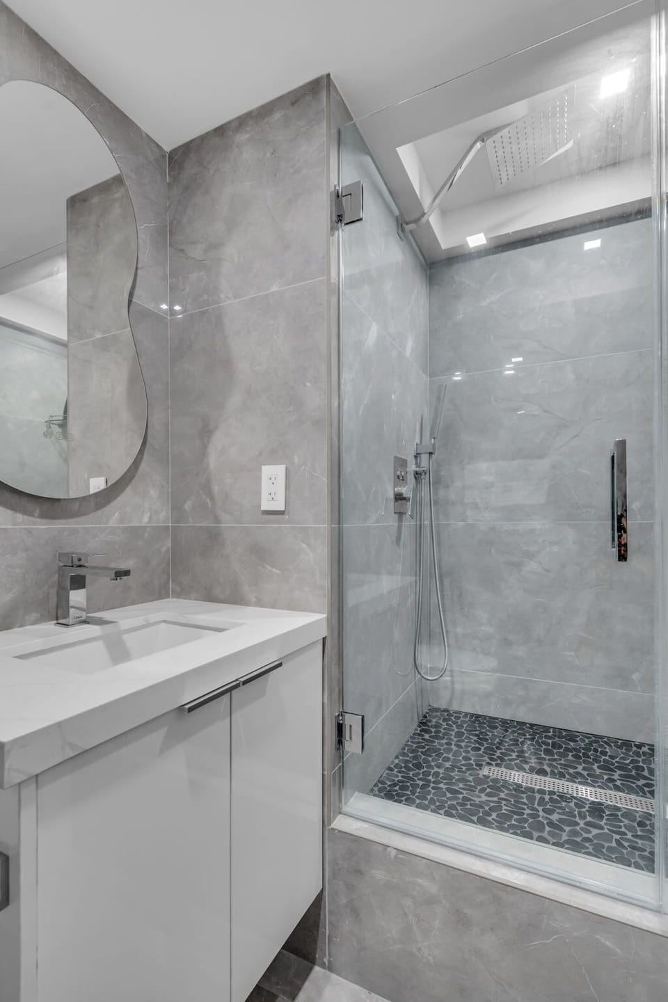 The modern guest bathroom features a sleek, glass-enclosed walk-in shower with a decorative pebble-tile floor. This bright space is completed with contemporary gray stone tiling and a clean white vanity.