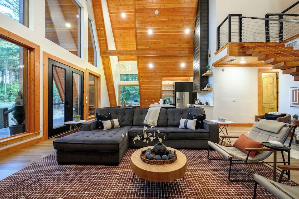Open concept living room with soaring wood ceilings, floor-to-ceiling windows, and loft staircase