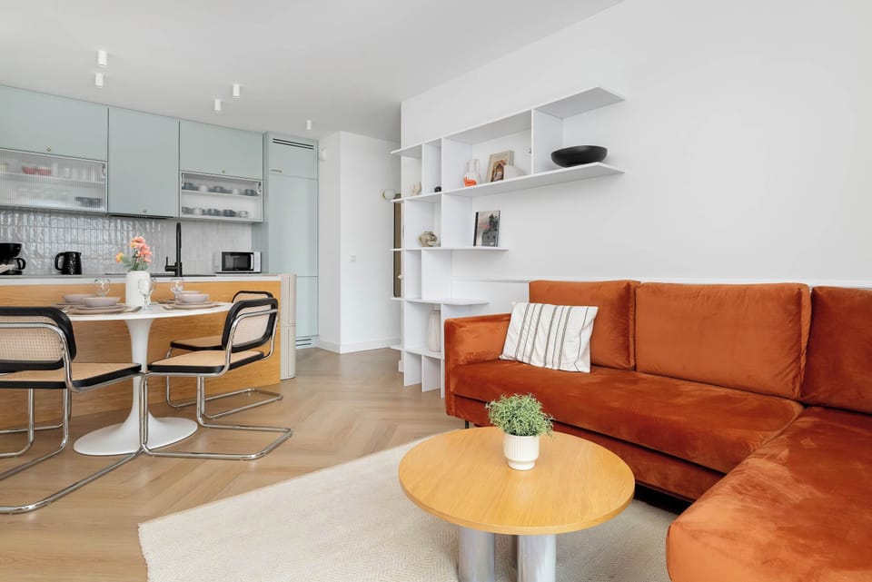 A spacious living room features a vibrant orange corner sofa and a wooden coffee table set on a light rug. Large windows and a dining area in the background create an open, airy atmosphere.